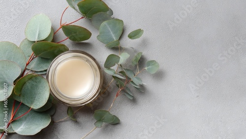 A small glass jar of pale cream lip balm sits on a grey surface, adorned with eucalyptus sprigs