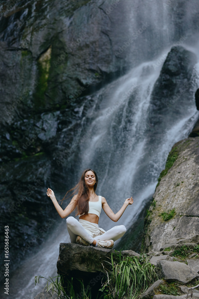 Fototapeta premium young woman meditating near waterfall in nature, peaceful, calm, yoga practice by rocks and flowing water with green plants