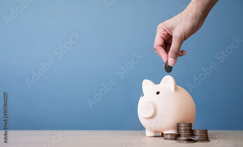Hand of woman putting coin into piggy bank on a wooden table with blue background. saving money and invest concept.