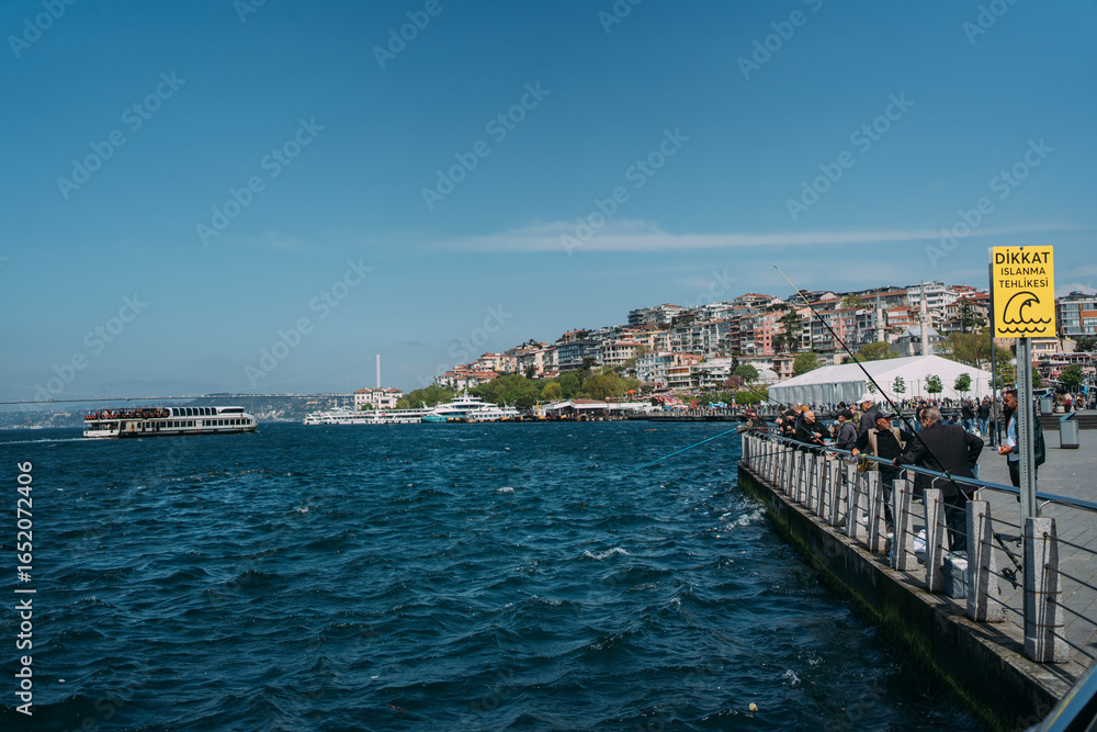 Naklejka premium Scenic waterfront promenade with people, boats, and a yellow Turkish warning sign by the blue sea under clear sky. Vibrant urban life, leisure, travel, and safety.