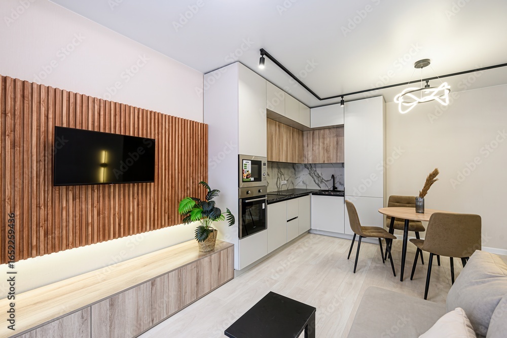 Fototapeta premium Modern apartment with TV on wood-paneled wall, sleek kitchen cabinets, and a cozy dining area. Neutral color palette with wood accents