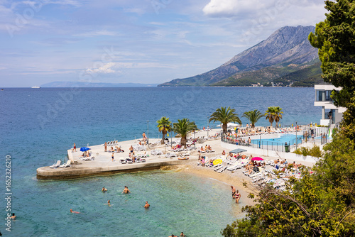 Fototapeta Naklejka Na Ścianę i Meble -  Beach with sun loungers and palm trees against the backdrop of the Adriatic Sea and mountains. Zaostrog village, Makarska Riviera, South Dalmatia, Croatia.