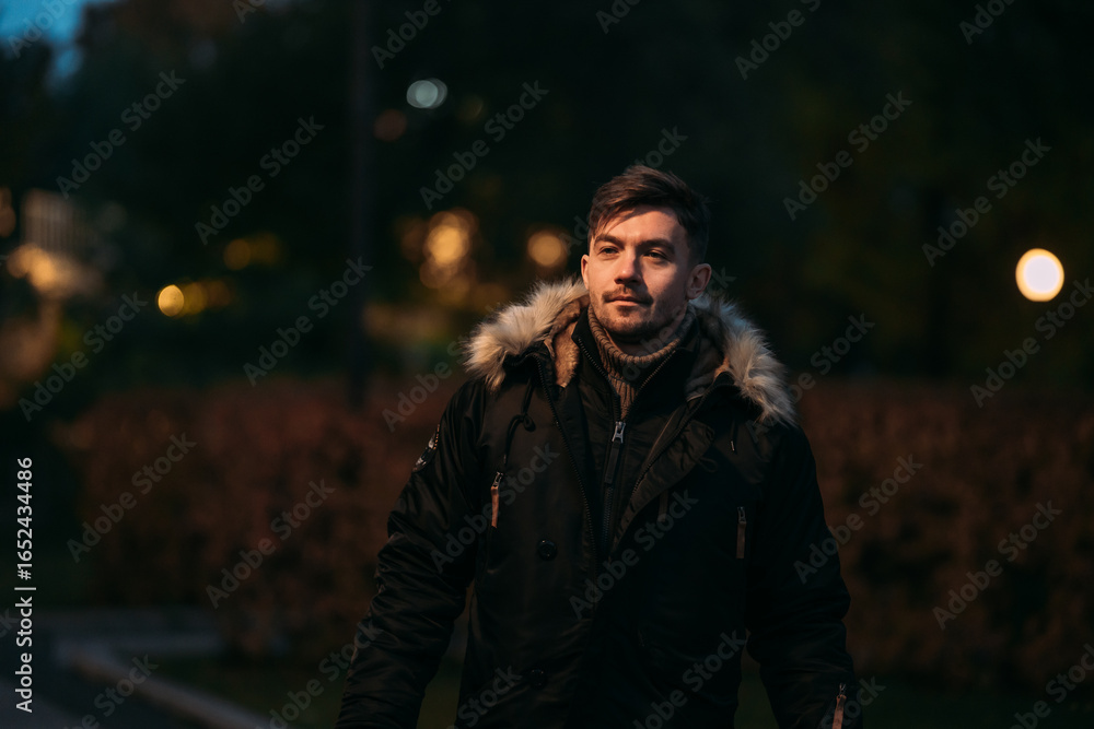 Obraz premium Person in black winter coat with fur hood standing outdoors in evening park. Warm clothing, cozy autumn atmosphere, street lights, modern fashion, and urban night stroll