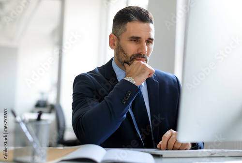 Concentrated middle aged male entrepreneur working with computer, looking at screen and thinking, focused businessman working in office interior, free space
