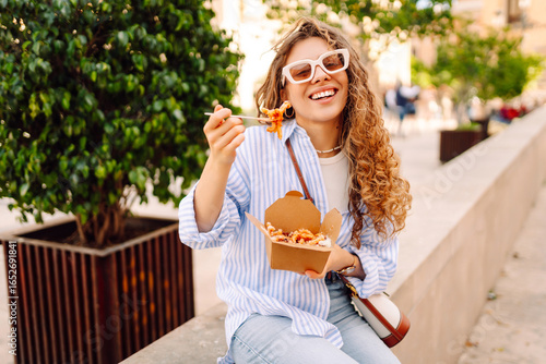 Fotografi Smiling tourist eats street food from a takeaway box on sunny city streets