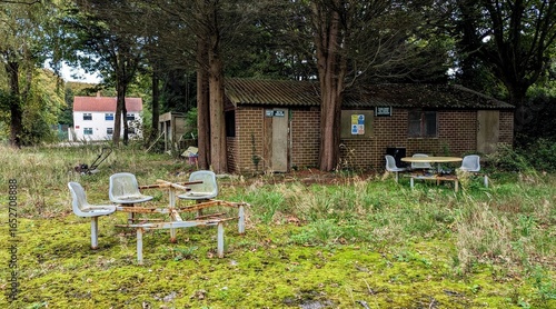 Post-apocalyptic park scene, rusty metal benches, overgrown grass, moss, forest trees, nature reclaiming. Weathered plastic chairs, picnic tables. Rusting outdoor seating. Deserted cabin wasteland.