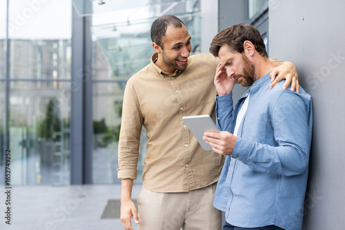 A supportive colleague comforts another while discussing something on a tablet in a modern office setting.