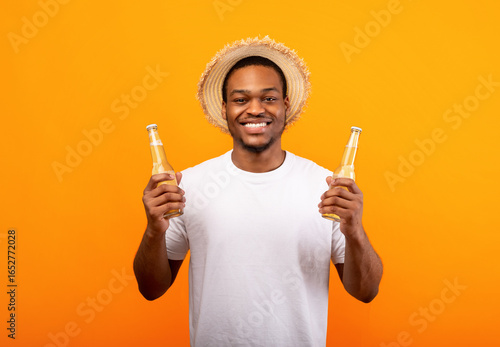 Canvas Print Cheerful young African American man holding two bottles of beer and smiling at camera on yellow studio background