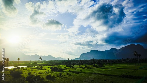 Sunrise over lush green fields in Aralvaimozhi, Kanyakumari, with dramatic clouds, distant mountains, and palm trees creating a stunning natural landscape