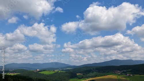 Hyperlapse aerial footage over the Bavarian Forest with a panoramic view towards the Czech Republic. Dramatic depth of slowly moving clouds across a blue and white sky above rolling hills, forests, an