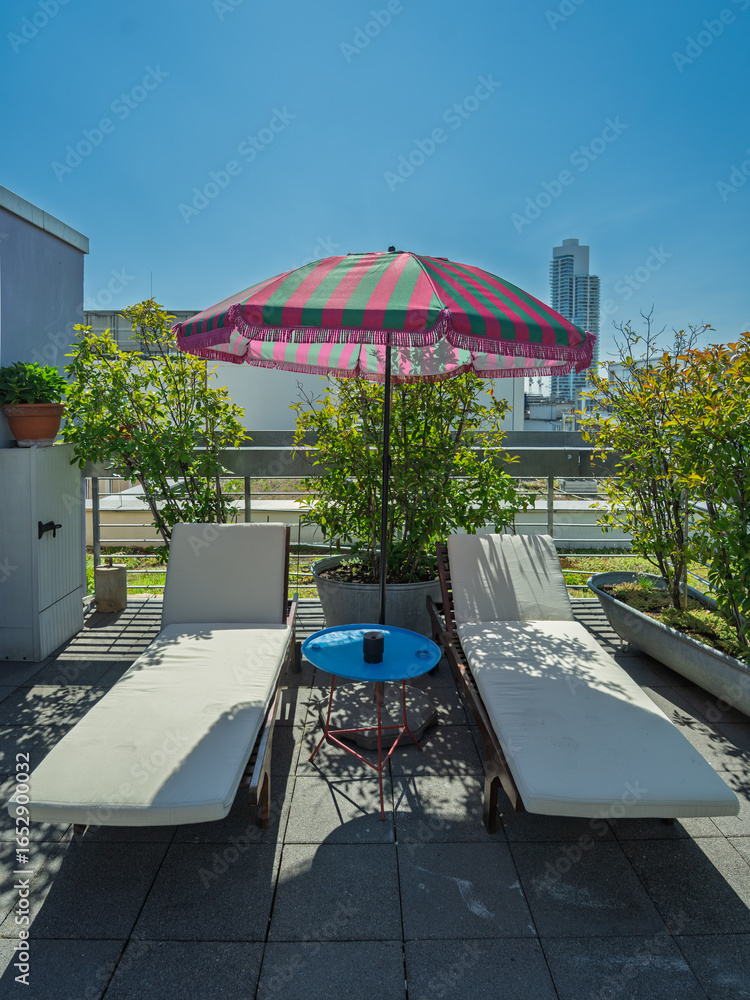 Fototapeta premium Two white sun loungers with a colorful striped umbrella and small blue table on a rooftop terrace with plants and a city skyline in the background