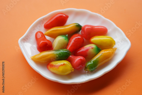 Colorful traditional Thai dessert called Luk Chup made from mung beans, coated with jelly, shaped into colorful miniature fruits, displayed on a white plate with bright orange background.  
