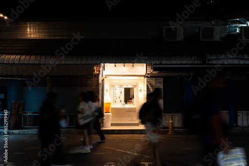 Small street-side shop warmly lit at night with motion-blurred pedestrians on an urban street