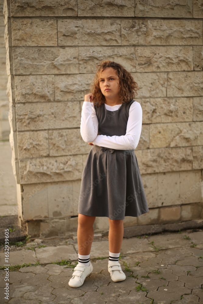 Portrait of a schoolgirl with curly hair wearing grey school uniform dress and white shirt outdoors near stone building, back to school concept Portrait of a schoolgirl with curly hair wearing grey school uniform dress and white shirt outdoors near stone building, back to school concept