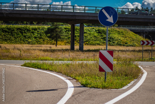 A curved road at a highway junction featuring directional traffic signs, including a blue arrow pointing down and a red-and-white chevron marker. An overpass bridge with a vehicle is visible in the ba