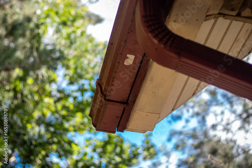 Close-up of a cabin roof corner showing a red rain gutter and downspout against a bright sky and leafy tree background.