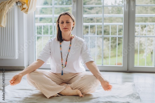 Woman practicing kundalini yoga at home, sitting in lotus pose with closed eyes and meditation beads. Spiritual practice, inner peace, and mindful coaching concept in natural light interior