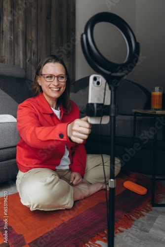 Smiling woman in glasses gives online consultation or remote lesson via smartphone and ring light. Home interior. Concept: video call, freelance, online learning, remote work