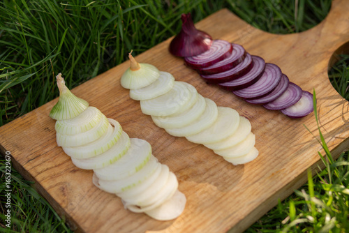 Close-up image of sliced yellow, red, and white onions arranged on a rustic wooden board placed on green grass, highlighting fresh farm produce, natural ingredients, and healthy cooking preparation