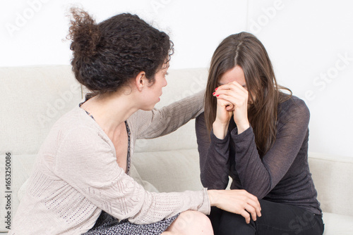 A young woman consoles her crying friend on the sofa, symbolizing therapy, emotional health, crisis support, compassion, and mental well-being
