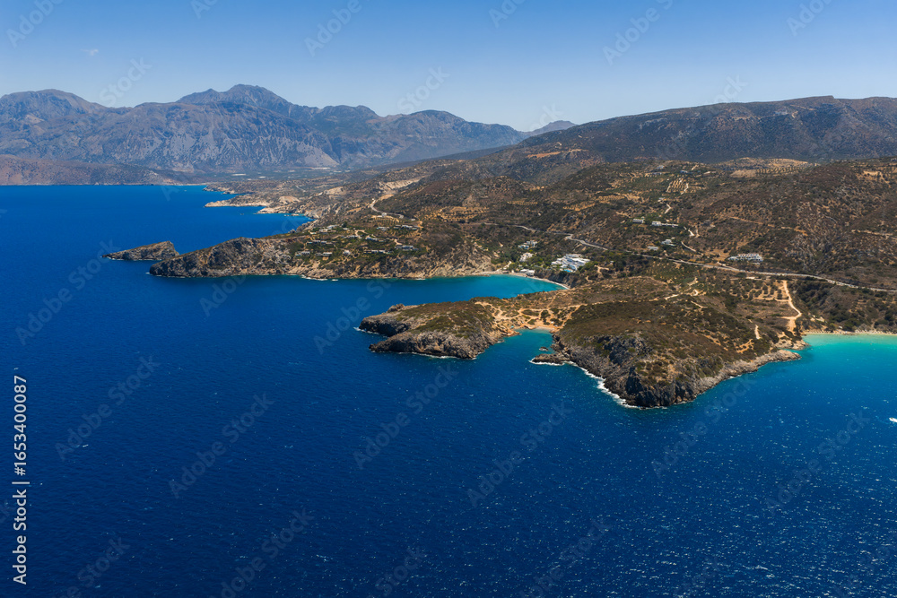 Obraz premium Aerial view of Voulisma Beach in Crete, Greece, featuring turquoise waters, golden sand, rugged coastline, arid hills, and distant mountains.