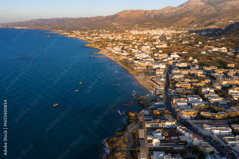 Fototapeta premium Aerial view of Stalida, Crete, showing clear blue waters, sandy beaches, small boats, white and beige buildings, and rugged sunlit mountains.