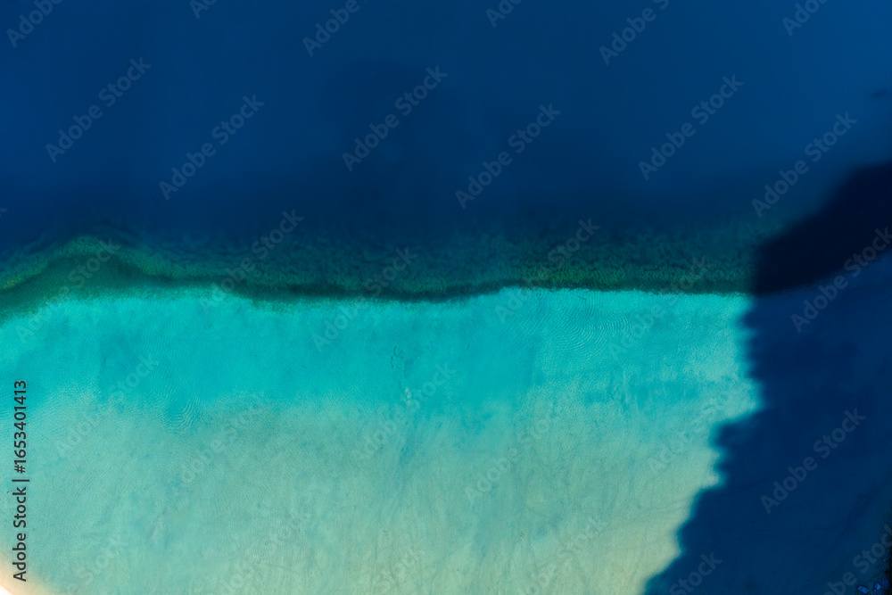 Fototapeta premium Aerial view of Lake Kournas in Crete, Greece, showing a gradient of turquoise to navy blue waters, visible sandy bottom, and contrasting shadows.