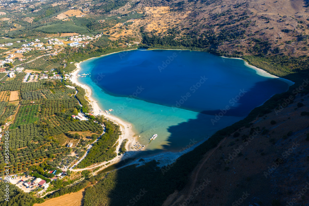 Obraz premium Aerial view of Lake Kournas in Crete, Greece, showing blue waters, sandy beaches, small boats, olive groves, arid hills, and nearby buildings.