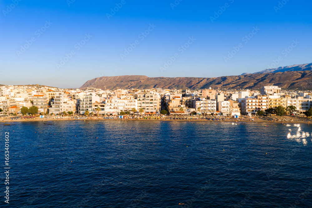 Chania town in Crete features white and pastel buildings by the sea, with rugged sunlit mountains in the background under a clear blue sky. Chania town in Crete features white and pastel buildings by the sea, with rugged sunlit mountains in the background under a clear blue sky.