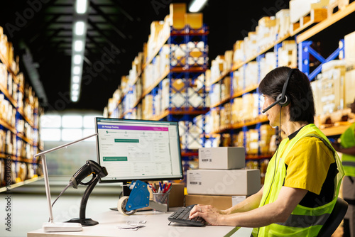 Asian worker in a hi vis vest answers a customer service call in a warehouse logistics center, assisting with tracking shipments, fixing a wrong address issue and updating shipping details.