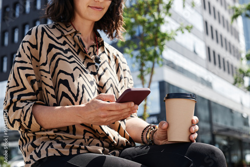 Horizontal medium section shot of unrecognizable young woman wearing stylish shirt sitting outdoors holding cup of coffee watching something in Internet on smartphone
