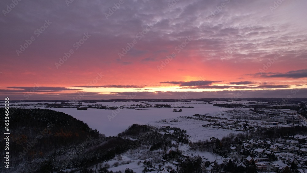 Fototapeta premium Panoramic view of a snow-covered landscape and frozen lake under a vivid winter sunset with dramatic purple and orange clouds.