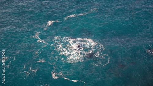 Looking down onto blue ocean waves washing over a rock in the middle of the sea.