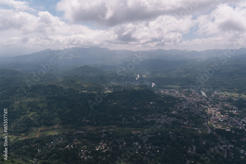 Aerial view of green tropical rainforest and river flowing through valley in Sri Lanka under cloudy sky, view from Ambuluwawa Tower