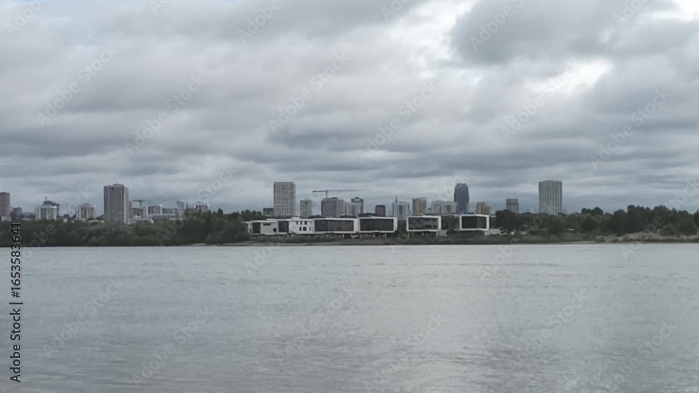 Naklejka premium View across a wide river to residential buildings on the opposite bank on a cloudy summer morning.