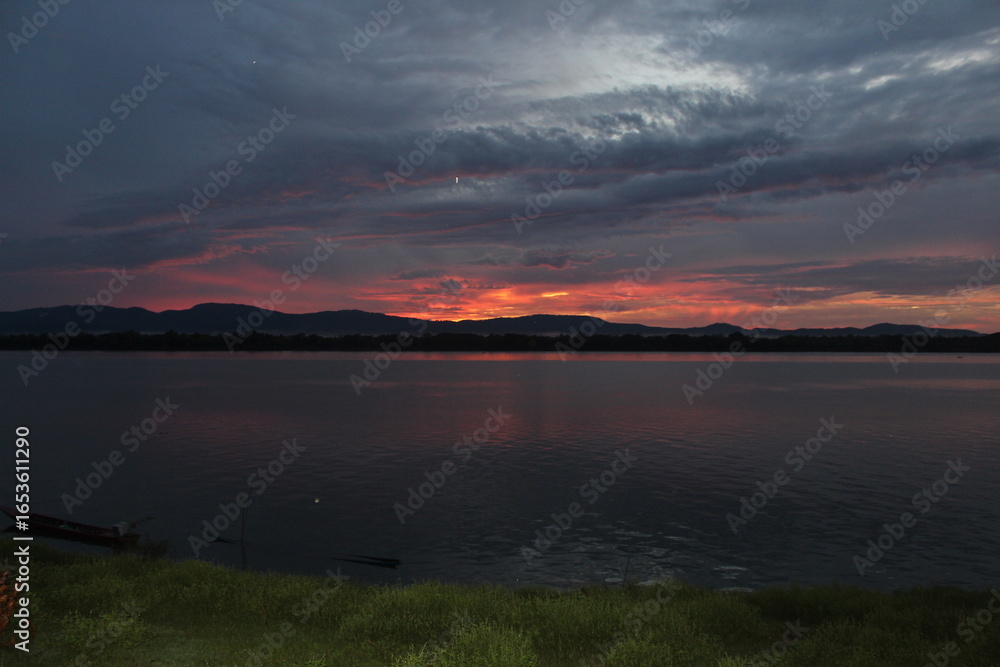 Fototapeta premium A breathtaking sunset over a calm river with vibrant orange and purple hues painting the sky. A small boat glides across the water, leaving gentle ripples under the silhouette of distant mountains.