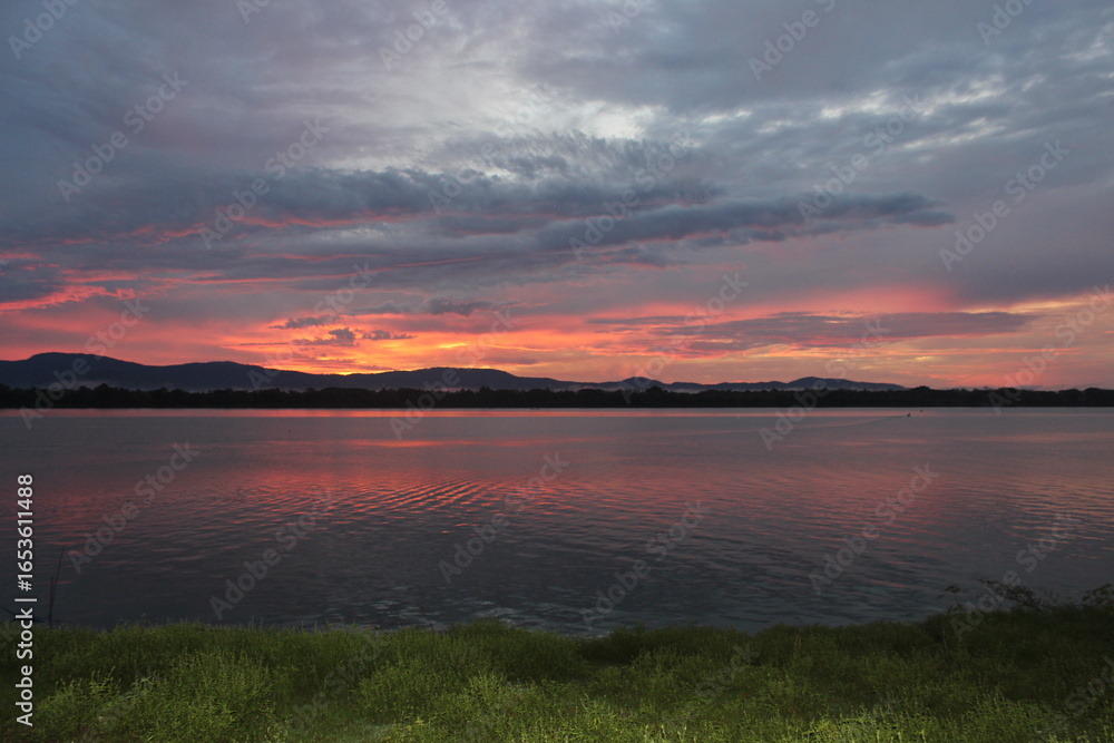Fototapeta premium A breathtaking sunset over a calm river with vibrant orange and purple hues painting the sky. A small boat glides across the water, leaving gentle ripples under the silhouette of distant mountains.