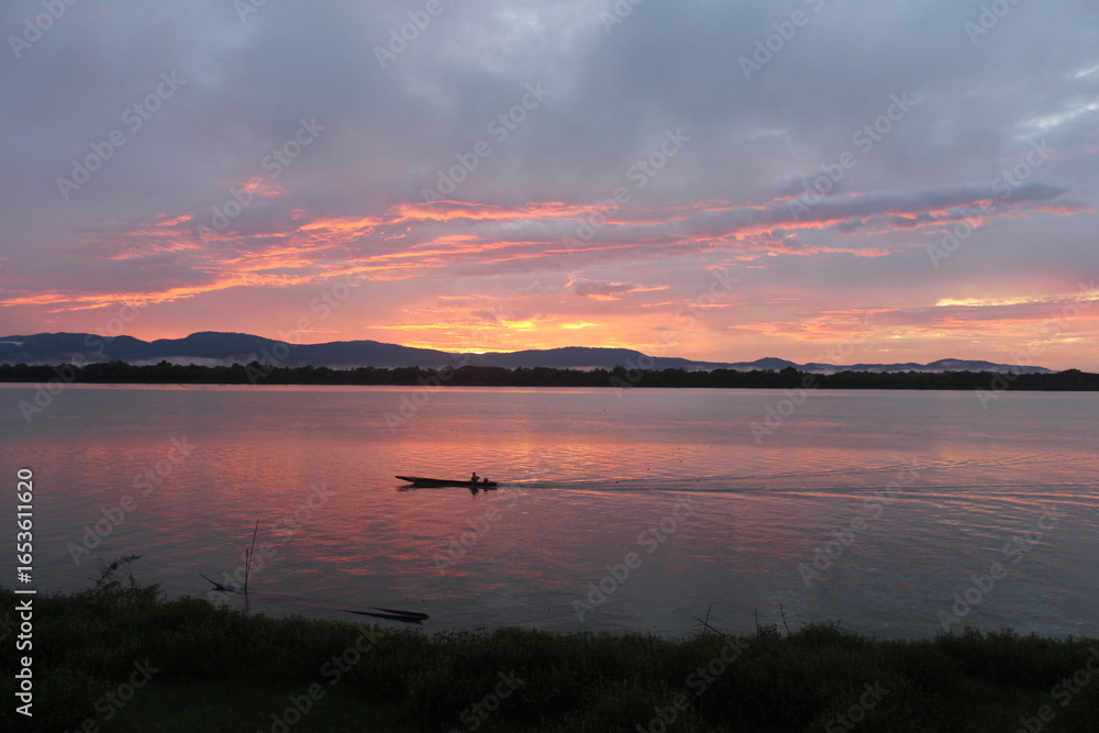 Fototapeta premium A breathtaking sunset over a calm river with vibrant orange and purple hues painting the sky. A small boat glides across the water, leaving gentle ripples under the silhouette of distant mountains.