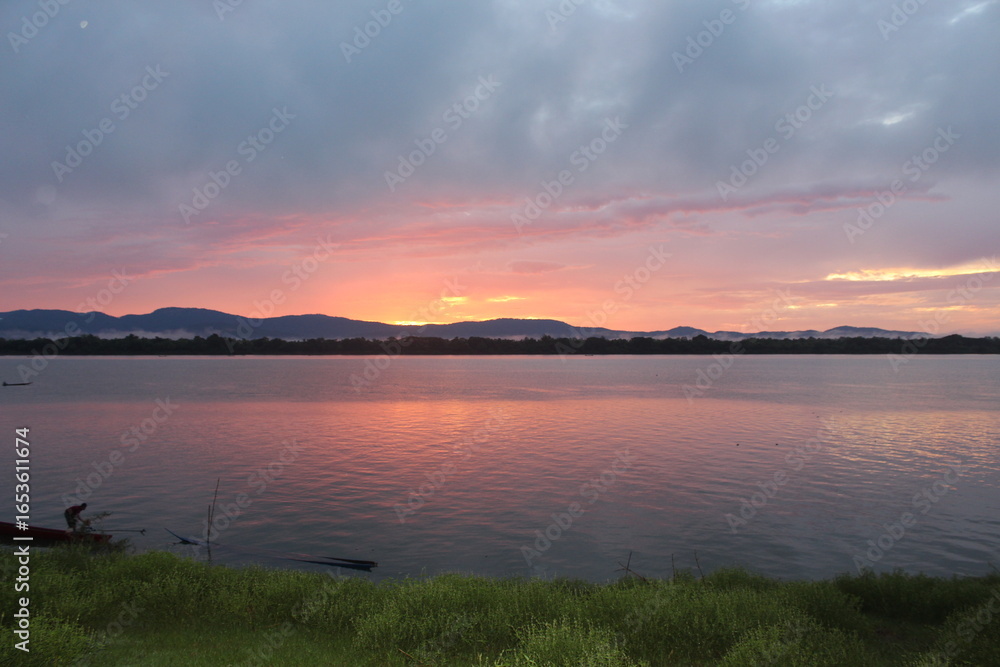Fototapeta premium A breathtaking sunset over a calm river with vibrant orange and purple hues painting the sky. A small boat glides across the water, leaving gentle ripples under the silhouette of distant mountains.