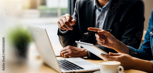 Two people discuss business at a desk with a laptop and documents, focusing on strategy planning and professional collaboration in a bright office environment.