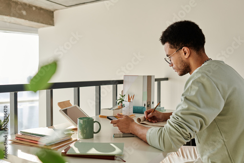 Horizontal image of college student sitting at desk and studying at home using digital tablet in his study