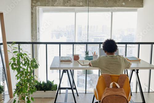 Horizontal rear view of little schoolboy sitting at his desk and studying with books in the modern room at home