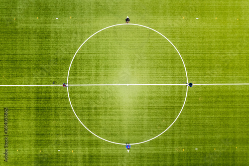 Aerial view of a vibrant green sports field marked with stark white lines and diminutive figures adding scale, HenningsvÃ¦r, Nordland, Norway.