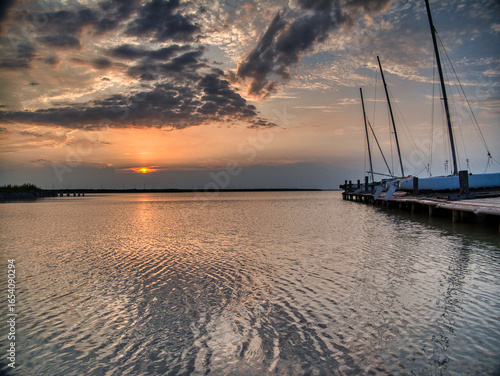 Peaceful sunrise at Breitenbrunn, Lake Neusiedl, Austria. Sailboats moored at a wooden pier with golden morning light reflecting on calm rippling water, symbolizing nature, sailing, and tranquility.
