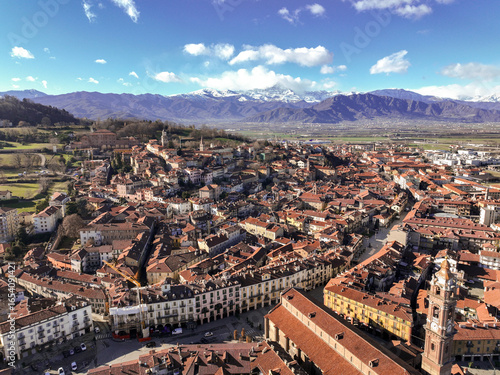 Aerial view of sun-kissed terracotta roofs cascade across the ancient town, nestled beneath the snow-capped Alps, revealing a timeless Italian vista, Saluzzo, Piedmont, Italy.