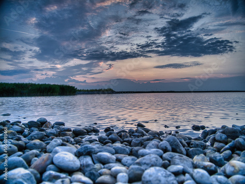 Peaceful blue hour sunrise at Breitenbrunn, Lake Neusiedl, Austria. Pebble shore, calm rippling water, and dramatic clouds create a tranquil lakeside scene, perfect for nature, travel, and landscape t