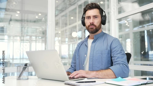 Wallpaper Mural Portrait of confident businessman in wireless headphones sitting at desk at workplace in modern business office. Successful handsome IT specialist busy with project working on laptop looking at camera Torontodigital.ca