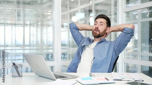 Happy businessman finished work on laptop while sitting at desk at workplace in a modern business office. Smiling satisfied man puts his hands behind his head, stretches himself in a chair. Work done