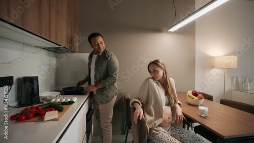 A man stands and cooks on a frying pan while talking to a pregnant woman sitting next to him on a chair
