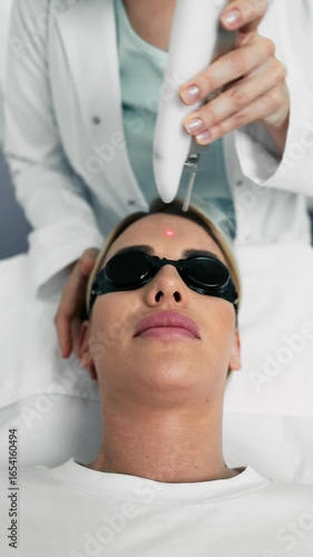 Beautician using laser device for removing facial wrinkles of female patient wearing protective glasses lying on medical couch in beauty salon
